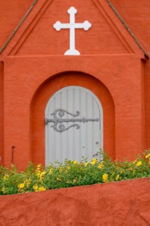 Detail of an orange-coloured church building with a white cross and Tor tor, yellow flowers in the