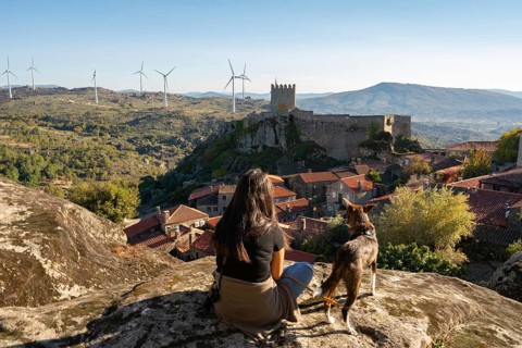 Woman and dog sit on a stone viewing of Sortelha castle and antique stone houses and wind turbines,