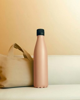 A pink water bottle with a black cap stands next to a beige bag on a beige couch with soft lighting