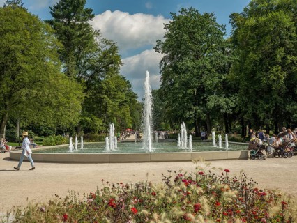 Central fountain in the park with walkers and lush greenery under a blue sky, Bad Lippspringe,