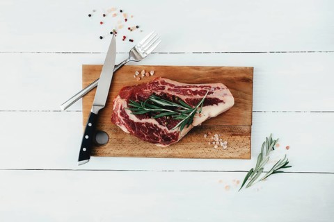 Raw ribeye steak on board, knife, fork, pepper, rosemary and pink Himalayan salt.