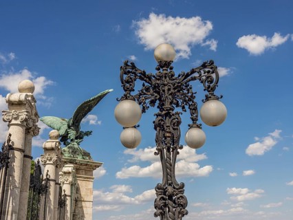 Wrought iron street lamp with ball lamps in front of a blue sky and a winged statue, budapest,