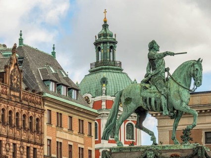 Bronze statue of a rider on a horse in front of a building with a central tower and clear sky,
