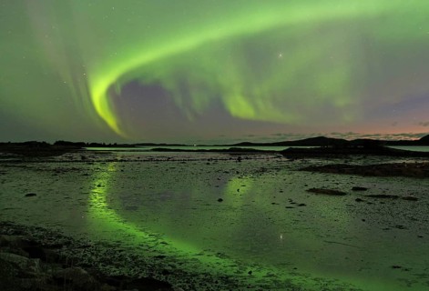 Northern lights (aurora borealis) cover the sky and are reflected in the water of a fjord,
