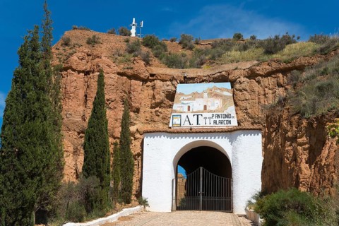 Entrance gate to a white building with a rock face and trees, surrounded by rustic charm on a sunny