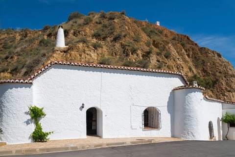 White-painted building with chimney and windows in front of a rock face, under a sunny blue sky,