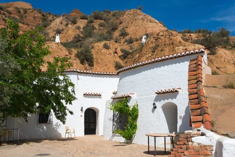 White building with rock wall, tree, and rustic outdoor furniture under a sunny blue sky, cave