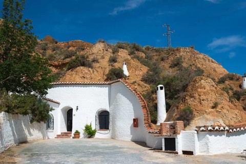 Small white building with chimney, tree and rock face in the background under a sunny blue sky,