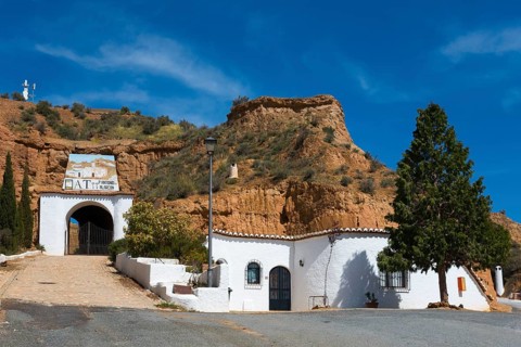 White building with entrance gate and trees in front of a rock face, under a sunny, cloudy sky,