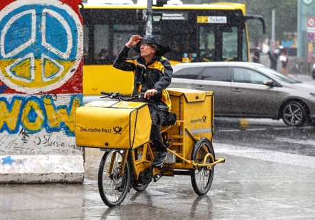 A DHL mail carrier rides his e-bike along a pavement in heavy rain, Berlin, 23 06 2023