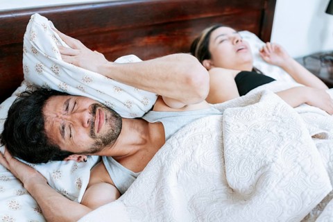 Woman snoring in bedroom and husband covering his ears. Husband suffering from the snoring of his