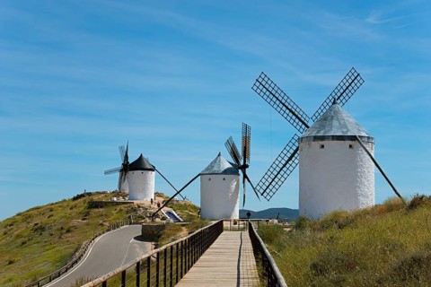 Wooden walkway leads to three white windmills on a green hill under a clear blue sky, Consuegra,