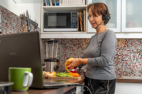 Woman, mid-50s, works from home, with laptop and communicates with colleagues via headset, home
