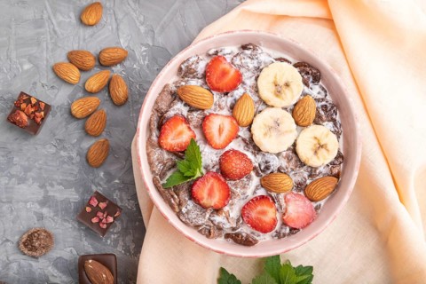 Chocolate cornflakes with milk, strawberry and almonds in ceramic bowl on gray concrete background
