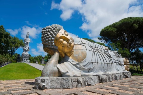 Monumental reclining Buddha statue in a green and peaceful park under a clear blue sky, Bacalhôa,