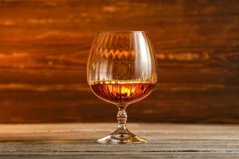 Round shiny glass filled with brandy or cognac, amber liquid on blurred wooden background