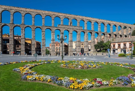A historic stone aqueduct with arches spans a city under a bright blue sky next to a colourful