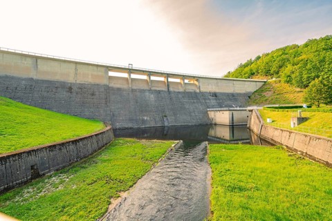 A mighty concrete dam with a river in the foreground, surrounded by green land and trees,