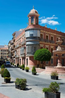 Historic building with bay window on an urban street with fountain and trees under a blue sky, Casa