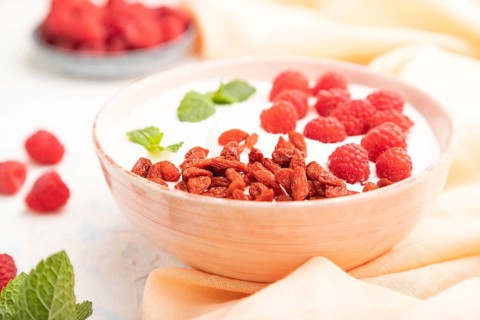 Yogurt with raspberry and goji berries in ceramic bowl on white concrete background and orange
