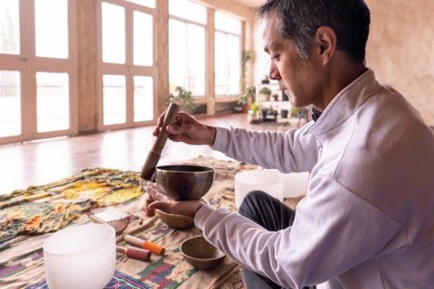 A middle-aged Asian man sits in profile, concentrating on gently vibrating a brass bowl in a softly