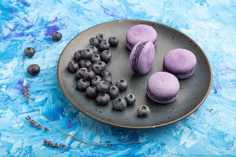 Purple macarons or macaroons cakes with blueberries on ceramic plate on a blue concrete background.
