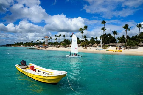 A sunny beach with clear blue water and a boat in the foreground, lighthouse, bar of the Hotel
