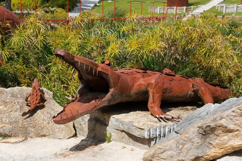 Metal sculpture of a crocodile with open mouth on a rock foundation surrounded by plants in the