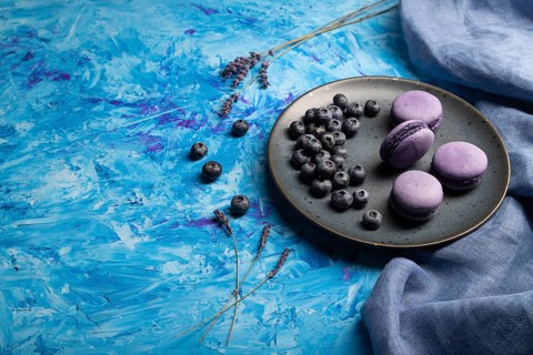Purple macarons or macaroons cakes with blueberries on ceramic plate on a blue concrete background