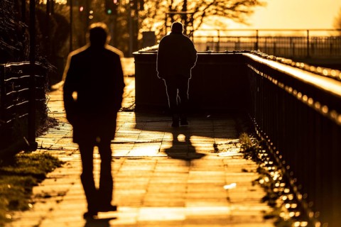 Man, elderly, walking on a pavement, a second man follows him, into the setting sun, long cast