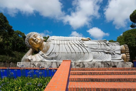 Reclining Buddha statue above an area decorated with flowers and steps in a sunny park, Bacalhôa,