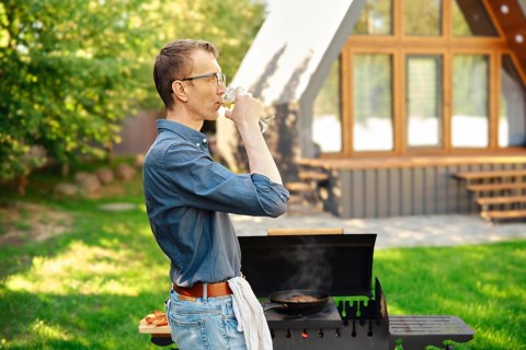 Middle-aged man drinks wine while standing in profile at the grill in the backyard of a log cabin