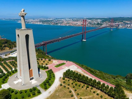 A tall statue with outstretched arms stands on a viewpoint near a bridge on the water, with a city