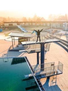 A person stretches his arms on a diving platform in an empty swimming pool in the sunlight, ENCW