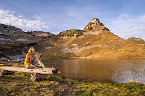 Lake Augstsee and the Atterkogel mountain on the Loser. A hiker sits on a bench and reads a book.