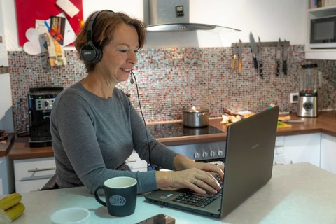 Woman, mid-50s, works from home, with laptop and communicates with colleagues via headset, home