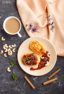 Cheese pancakes with caramel sauce on a beige ceramic plate and a cup of coffee on a black concrete