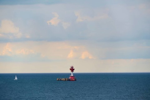 A red lighthouse stands on a calm, blue sea under a slightly cloudy sky, with a small sailing boat