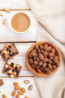 Almond in chocolate dragees in wooden plate and a cup of coffee on white wooden background and