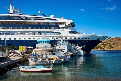 A large cruise ship moored in the harbour of a Greek island, surrounded by smaller fishing boats on