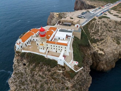 A red lighthouse on a cliff, surrounded by white buildings and a wide view of the ocean and the