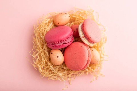 Purple and pink macaroons and chocolate eggs in decorative paper nest on pink pastel background.