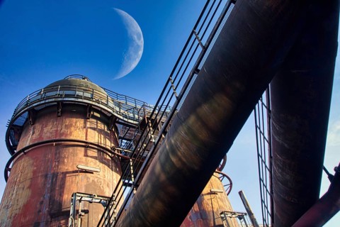 Rusted industrial plant in front of a clear blue sky with visible moon in the background,