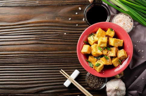 Flat lay view at stir fried tofu cubes with chives in clay dish on wooden kitchen table with napkin