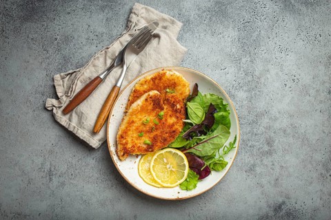 Crispy panko breaded fried chicken fillet with green salad and lemon on plate with cutlery on gray