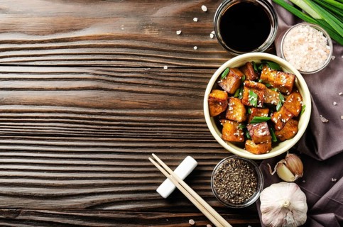 Flat lay view at stir fried tofu cubes with chives in clay dish on wooden kitchen table with napkin