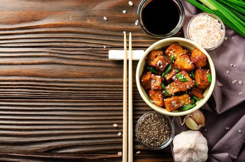 Flat lay view at stir fried tofu cubes with chives in clay dish on wooden kitchen table with napkin