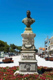 Stone bust on a pedestal with flowerbed in the foreground and clear sky in the background, monument