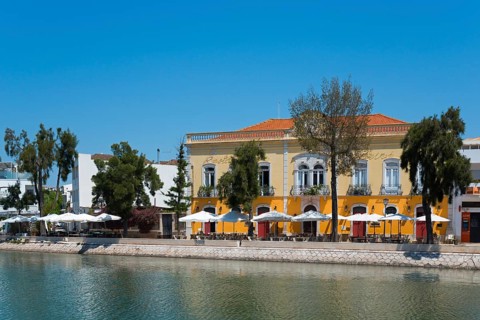 Yellow building with terrace area by the river, surrounded by trees and a restaurant in the