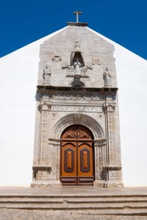 Historic church façade with detailed stonework and wooden door under a blue sky, Igreja da